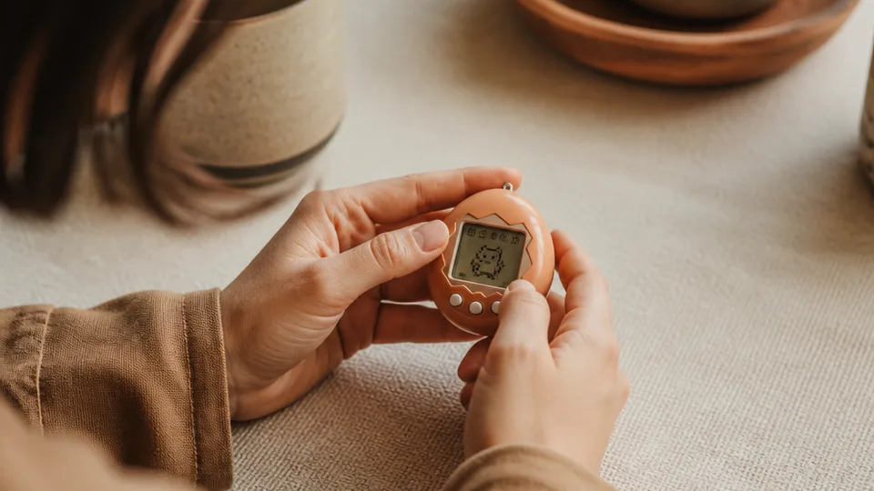 A person's hands holding a small handheld digital device, the tiny screen displaying a simple animated character that evokes real caregiving instincts